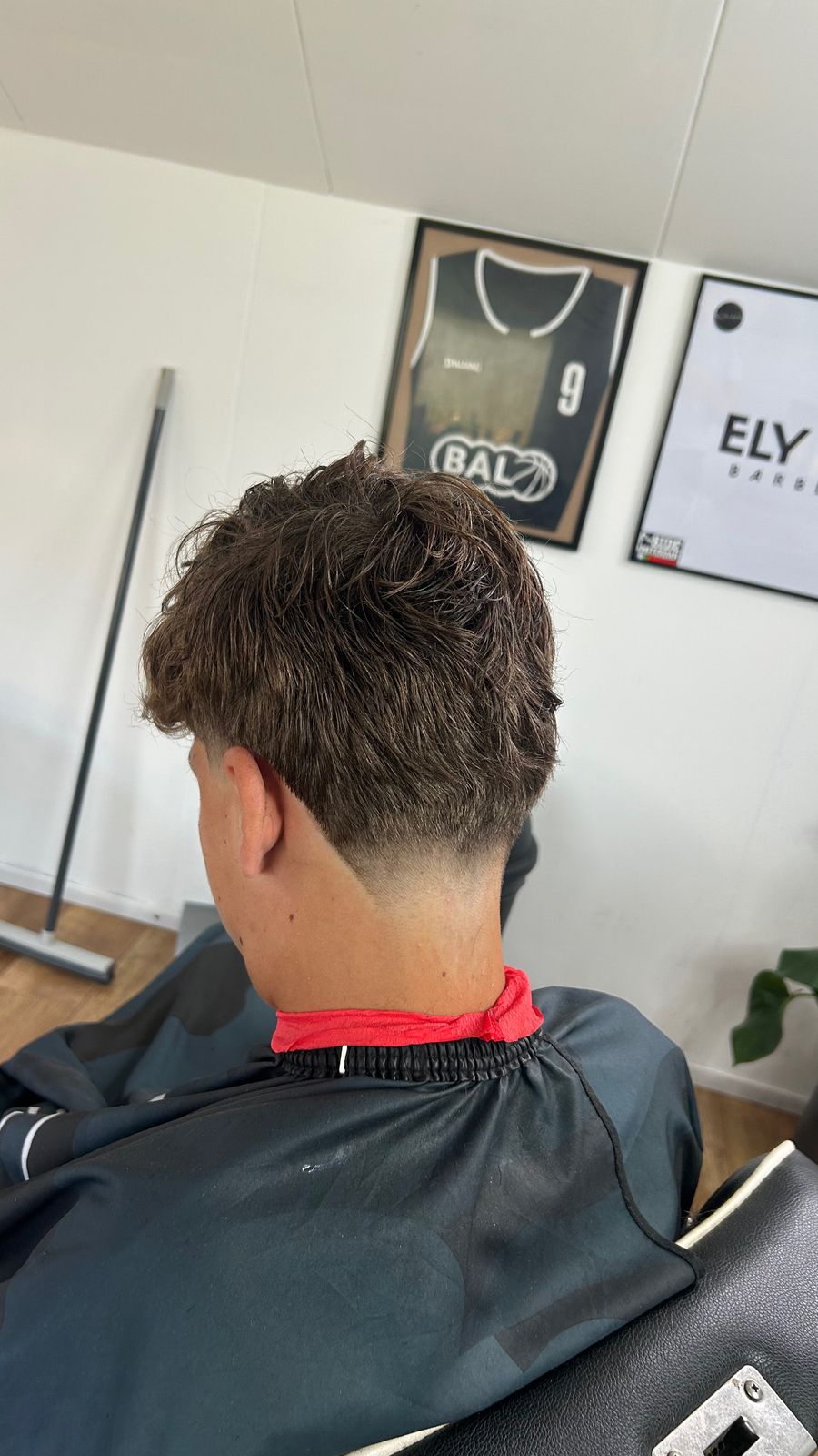 Achterhoofd van een klant met een verse fade, met het ELY-bord en een ingelijst basketbalshirt aan de muur